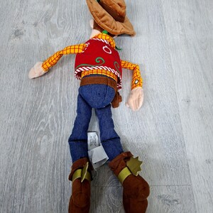 May include: A soft toy cowboy character, with a brown hat, red vest with white detailing, yellow plaid shirt, blue jeans, and brown boots with gold accents. The toy is on a light grey wooden surface.