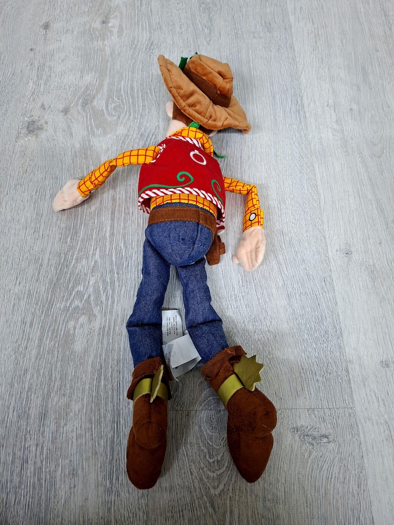May include: A soft toy cowboy character, with a brown hat, red vest with white detailing, yellow plaid shirt, blue jeans, and brown boots with gold accents. The toy is on a light grey wooden surface.