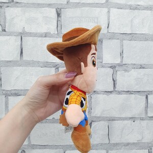 K&ouml;nnte beinhalten: Ein Pl&uuml;schtier der Figur Woody aus dem Toy Story-Franchise. Die Puppe tr&auml;gt einen braunen Cowboyhut, ein gelbes Hemd mit schwarzen Kuhflecken, ein rotes Halstuch und eine blaue Hose. Das Spielzeug ist aus weichem Stoff.