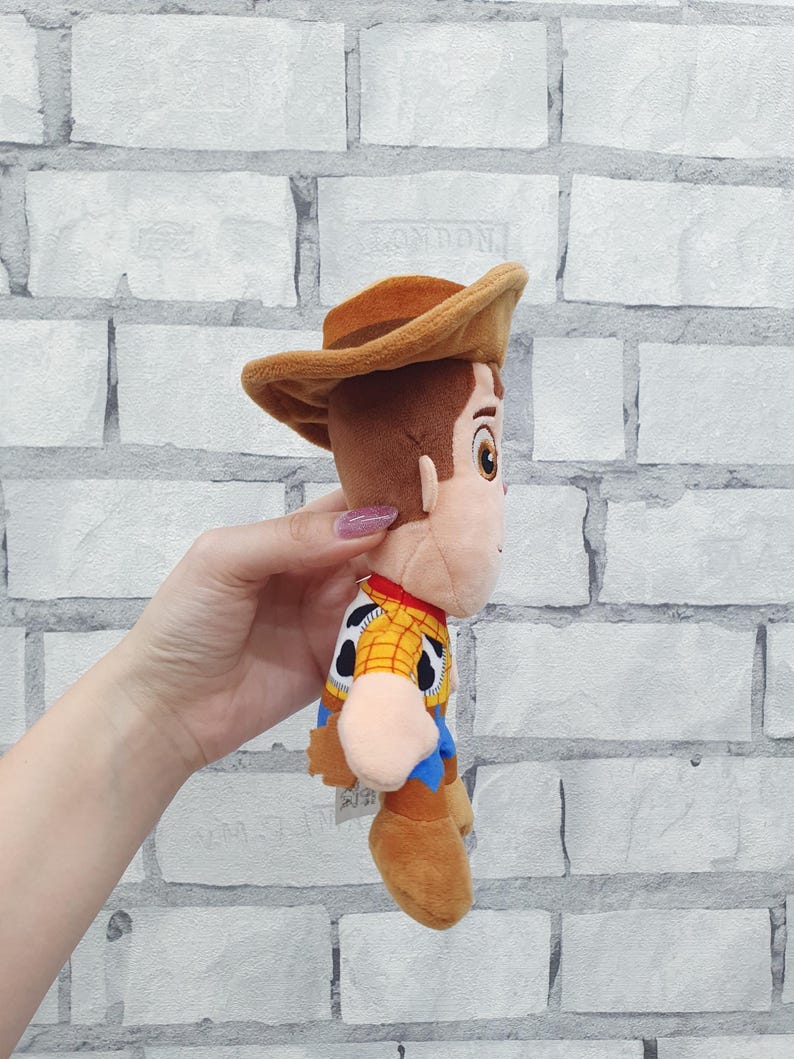K&ouml;nnte beinhalten: Ein Pl&uuml;schtier der Figur Woody aus dem Toy Story-Franchise. Die Puppe tr&auml;gt einen braunen Cowboyhut, ein gelbes Hemd mit schwarzen Kuhflecken, ein rotes Halstuch und eine blaue Hose. Das Spielzeug ist aus weichem Stoff.