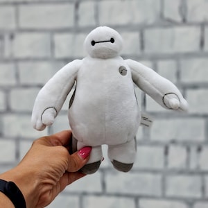 Può includere: Un peluche bianco con una linea nera per gli occhi e un piccolo cerchio nero sul petto. Le braccia e le gambe sono arrotondate e attaccate al corpo. Il giocattolo è tenuto in una mano.