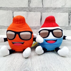 May include: Two plush toy figures, one orange and one blue, each with sunglasses, a smiling mouth, and white hands and feet. The orange figure has a red top, while the blue figure has a red and white top. Both are sitting.