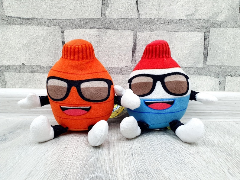 May include: Two plush toy figures, one orange and one blue, each with sunglasses, a smiling mouth, and white hands and feet. The orange figure has a red top, while the blue figure has a red and white top. Both are sitting.