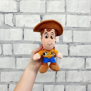 K&ouml;nnte beinhalten: Ein Pl&uuml;schtier eines Cartoon-Cowboy-Charakters, der einen braunen Hut, ein gelbes kariertes Hemd, eine blaue Hose und braune Stiefel tr&auml;gt. Das Spielzeug hat einen freundlichen Ausdruck und wird vor einer wei&szlig;en Backsteinmauer gehalten.