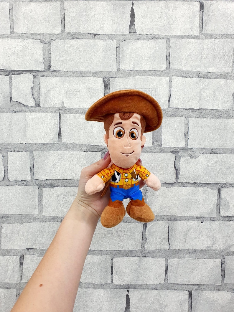 K&ouml;nnte beinhalten: Ein Pl&uuml;schtier eines Cartoon-Cowboy-Charakters, der einen braunen Hut, ein gelbes kariertes Hemd, eine blaue Hose und braune Stiefel tr&auml;gt. Das Spielzeug hat einen freundlichen Ausdruck und wird vor einer wei&szlig;en Backsteinmauer gehalten.