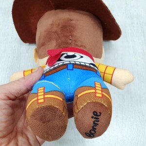 May include: A plush toy cowboy character, seen from behind. The toy features a brown cowboy hat, a red bandana, and blue trousers. The toy's boots are brown, and the word "Bonnie" is written on one of the boots.