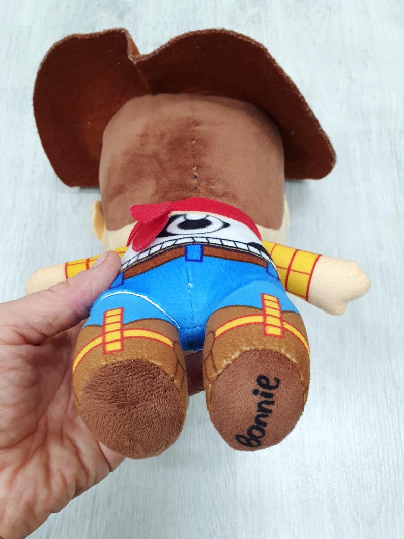 May include: A plush toy cowboy character, seen from behind. The toy features a brown cowboy hat, a red bandana, and blue trousers. The toy's boots are brown, and the word "Bonnie" is written on one of the boots.