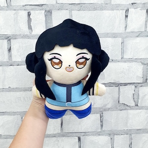 May include: A plush doll with a cartoon-style face, black hair, and a blue and white outfit. The doll has large, expressive eyes and a small, smiling mouth. The doll is held against a white brick wall.