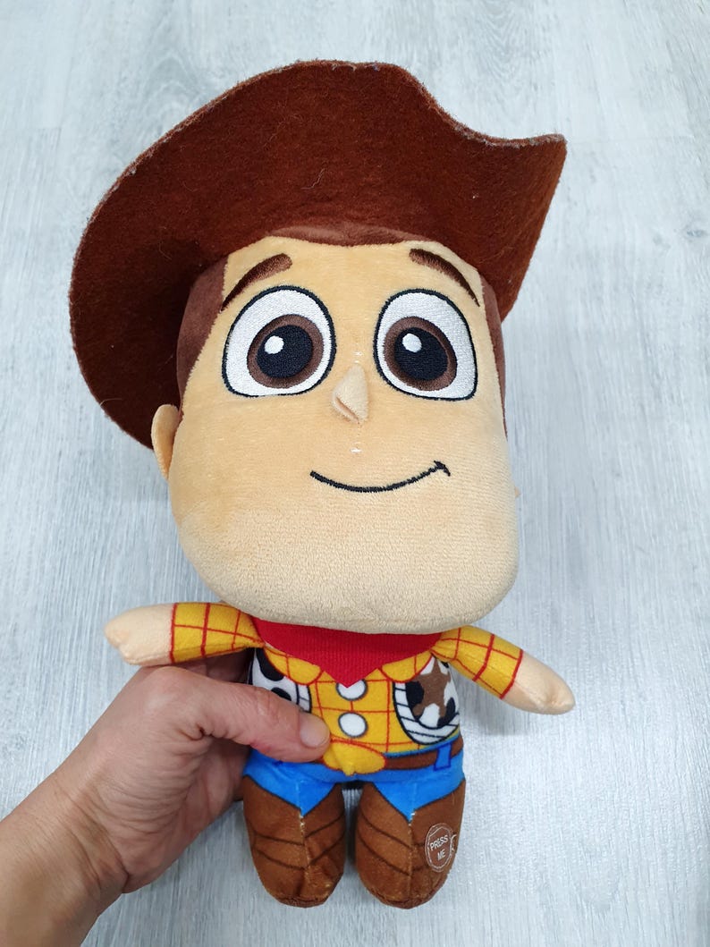 May include: A soft toy of the character Woody from Toy Story. The toy features a brown cowboy hat, a tan face, and a yellow shirt with a red neckerchief. It is wearing blue trousers and brown boots.