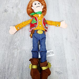 May include: A soft toy cowboy character, featuring a brown hat, yellow plaid shirt, red vest with a Christmas tree design, blue jeans, and brown boots with gold spurs. The toy has a green neckerchief and a gold belt buckle.