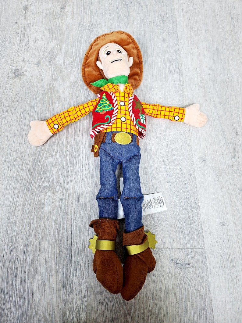 May include: A soft toy cowboy character, featuring a brown hat, yellow plaid shirt, red vest with a Christmas tree design, blue jeans, and brown boots with gold spurs. The toy has a green neckerchief and a gold belt buckle.