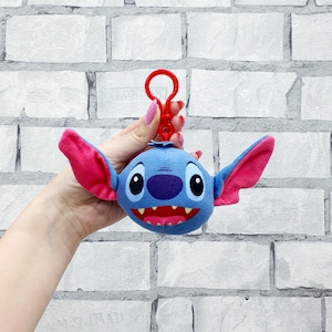 Może przedstawiać: Niebieski pluszowy brelok do kluczy Stitch z dużymi r&oacute;żowymi uszami i czerwonymi ustami. Brelok ma czerwony zatrzask. Pluszowa zabawka to postać z filmu Disneya Lilo & Stitch.
