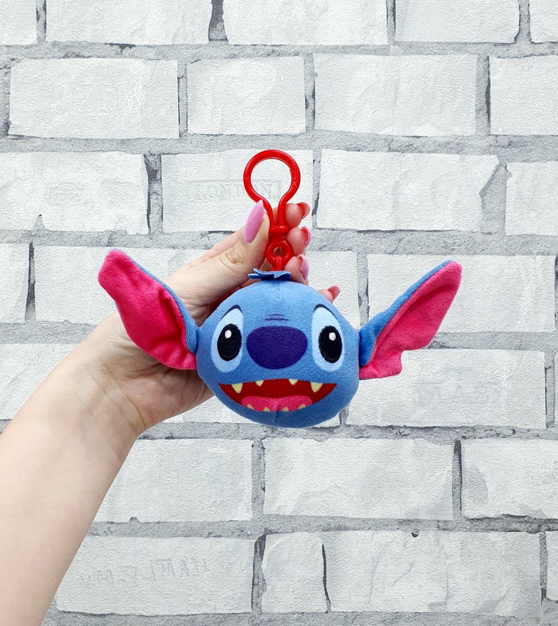 Może przedstawiać: Niebieski pluszowy brelok do kluczy Stitch z dużymi r&oacute;żowymi uszami i czerwonymi ustami. Brelok ma czerwony zatrzask. Pluszowa zabawka to postać z filmu Disneya Lilo & Stitch.