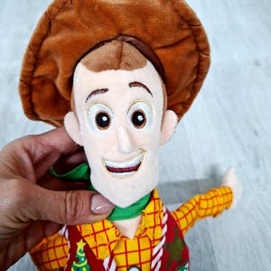 May include: A soft toy of a cartoon character wearing a brown cowboy hat, a yellow plaid shirt, and a red waistcoat with a Christmas tree design. The character has a green neckerchief and a smiling face.