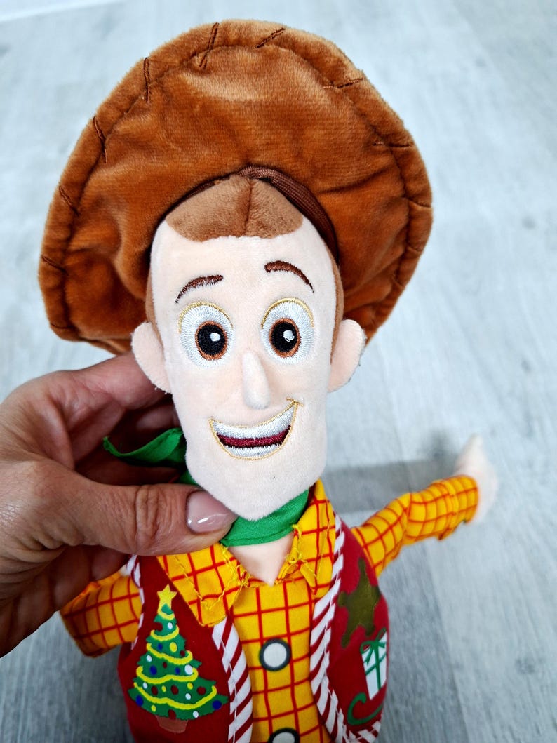 May include: A soft toy of a cartoon character wearing a brown cowboy hat, a yellow plaid shirt, and a red waistcoat with a Christmas tree design. The character has a green neckerchief and a smiling face.