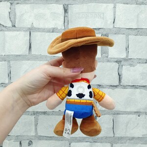K&ouml;nnte beinhalten: Ein Pl&uuml;schtier eines Cowboy-Charakters mit braunem Hut, gelbem kariertem Hemd, blauer Hose und braunen Stiefeln. Das Spielzeug hat ein wei&szlig;es Abzeichen mit einem schwarz-wei&szlig;en Design auf dem R&uuml;cken.