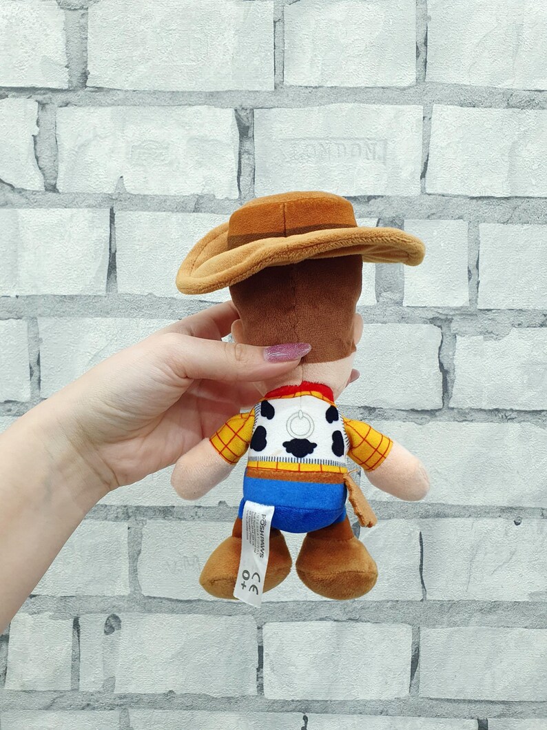 K&ouml;nnte beinhalten: Ein Pl&uuml;schtier eines Cowboy-Charakters mit braunem Hut, gelbem kariertem Hemd, blauer Hose und braunen Stiefeln. Das Spielzeug hat ein wei&szlig;es Abzeichen mit einem schwarz-wei&szlig;en Design auf dem R&uuml;cken.