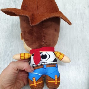 May include: A soft toy cowboy character with a brown hat, brown hair, and a red neckerchief. The toy is dressed in a white shirt, black vest, blue trousers, and brown boots. The arms are yellow with a checkered pattern.