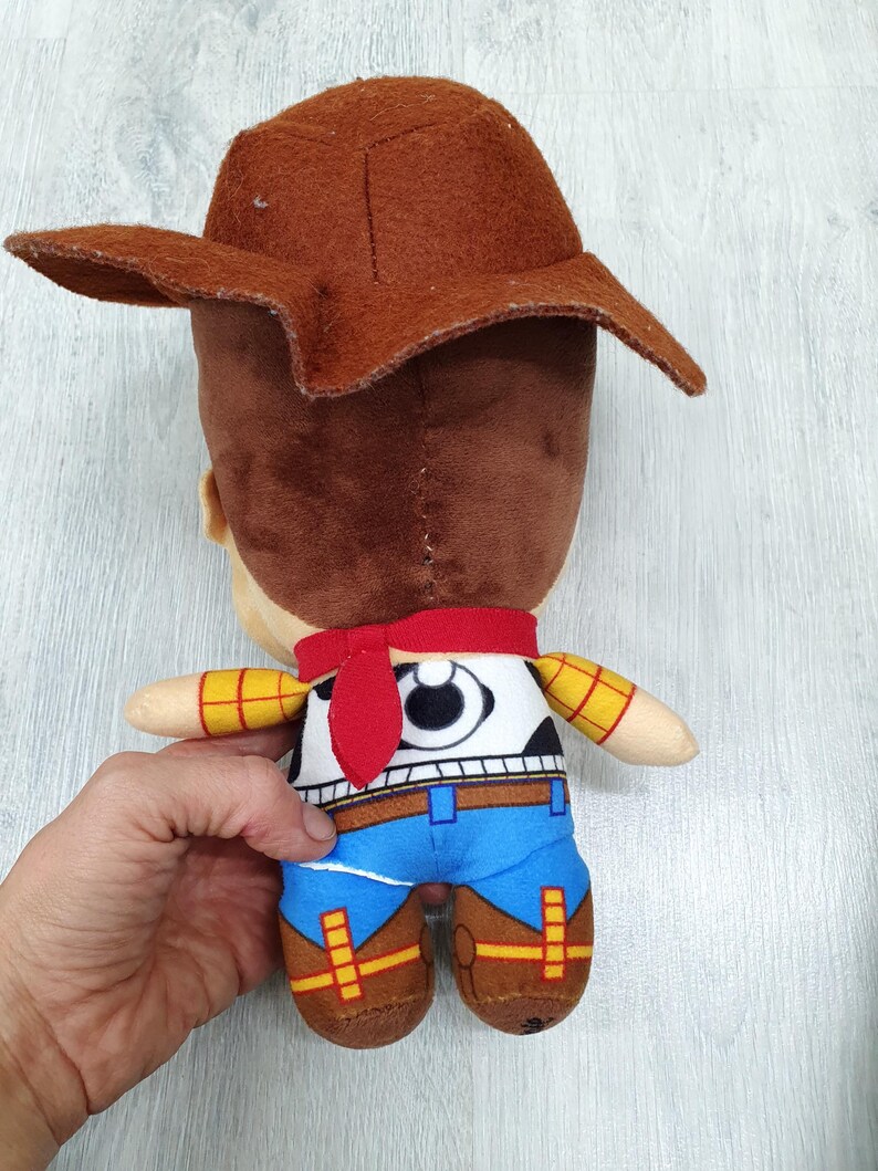 May include: A soft toy cowboy character with a brown hat, brown hair, and a red neckerchief. The toy is dressed in a white shirt, black vest, blue trousers, and brown boots. The arms are yellow with a checkered pattern.