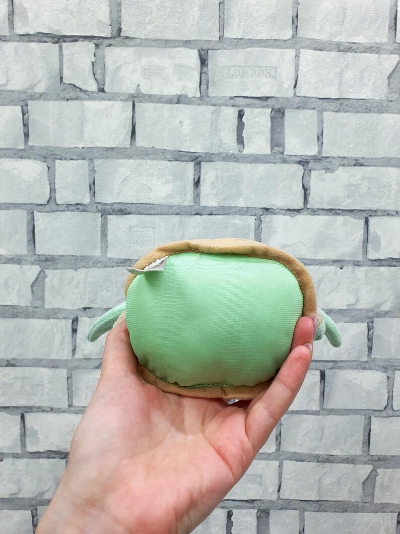 May include: A plush turtle toy in shades of green and tan, held in a hand. The turtle has a rounded green body, a tan shell, and small flipper-like appendages. A small tag is attached to the toy.