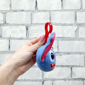 Może przedstawiać: Niebieska pluszowa postać z r&oacute;żowymi akcentami, czerwonymi ustami i czarnym okiem. Pluszowa zabawka ma przymocowany czerwony plastikowy klips. Tło to biała ceglana ściana.