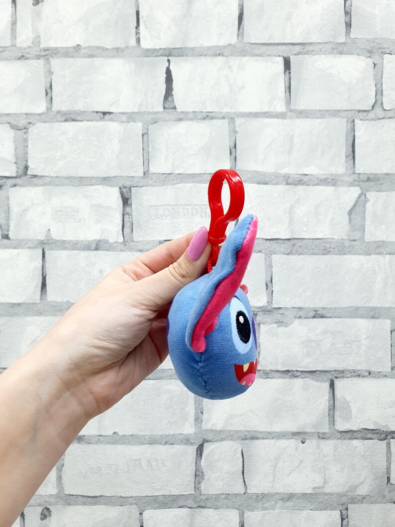 Może przedstawiać: Niebieska pluszowa postać z r&oacute;żowymi akcentami, czerwonymi ustami i czarnym okiem. Pluszowa zabawka ma przymocowany czerwony plastikowy klips. Tło to biała ceglana ściana.