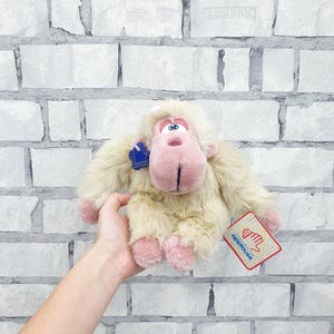 May include: A plush toy monkey with cream-colored fur, pink hands and feet, and a pink face. The monkey has a blue tag and a tag that says "applause". The toy is held up against a white brick wall.