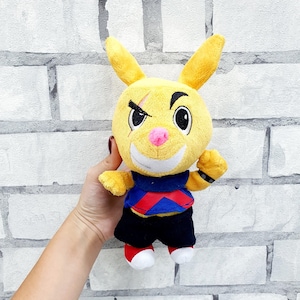 May include: A yellow plush rabbit toy with a black eye patch and a scar. The rabbit wears a blue, red, and black outfit. The toy has a pink nose and white teeth. The rabbit's feet are red and white. The toy is held in a hand.