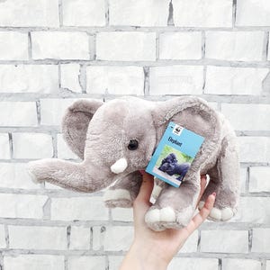 May include: A plush gray elephant toy with large ears, a trunk, and small tusks. The toy is held in a hand, and a blue tag with the word "Elephant" is attached. The background is a white brick wall.