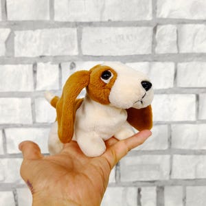 Può includere: Un peluche a forma di cane basset hound con pelo marrone e bianco, lunghe orecchie flosce e occhi neri. Il peluche è tenuto in mano su uno sfondo di muro di mattoni bianchi.