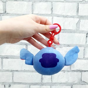 Może przedstawiać: Pluszowa niebieska postać z dużymi uszami i ciemnofioletową łatą na ciele, przymocowana do czerwonego plastikowego klipsa. Tło to biała ceglana ściana.