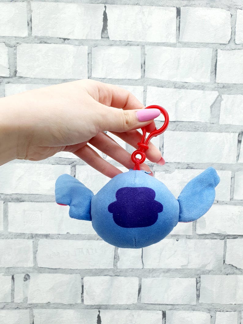 Może przedstawiać: Pluszowa niebieska postać z dużymi uszami i ciemnofioletową łatą na ciele, przymocowana do czerwonego plastikowego klipsa. Tło to biała ceglana ściana.