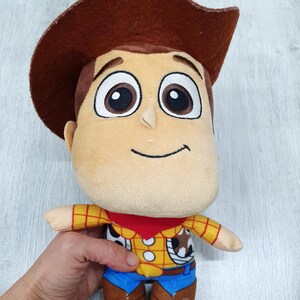 May include: A soft toy of the character Woody from Toy Story. The doll features a brown cowboy hat, a tan face, and a yellow shirt with a red neckerchief. Blue trousers and brown boots complete the outfit.