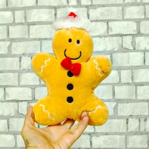 May include: A soft gingerbread man toy, wearing a white-trimmed Santa hat and a red bow tie. The yellow figure has a smiling face with black eyes and a mouth, and three black buttons. White zig-zag stitching outlines the arms and legs.