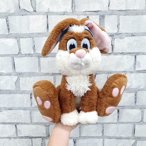 Puede incluir: Un peluche de conejo marrón con grandes ojos azules, orejas internas rosas y pecho blanco. El conejo tiene las patas extendidas con almohadillas rosas. Se sostiene contra una pared de ladrillos blancos.