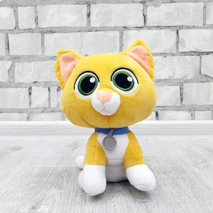 May include: A plush toy cat with yellow fur, white paws, and a white belly. The cat has large green eyes, a pink nose, and a blue collar with a gray tag. The cat is sitting on a light-colored wooden surface.