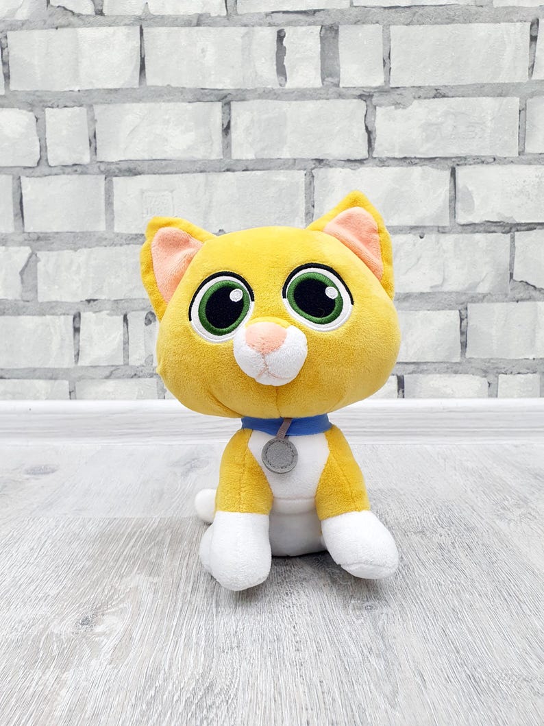 May include: A plush toy cat with yellow fur, white paws, and a white belly. The cat has large green eyes, a pink nose, and a blue collar with a gray tag. The cat is sitting on a light-colored wooden surface.