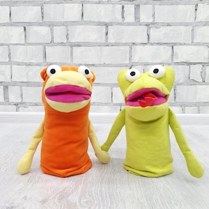May include: Two handmade puppet toys. One is orange with a yellow face and pink lips, the other is green with a yellow face and pink lips. Both have black eyes and small arms. They are standing side by side.