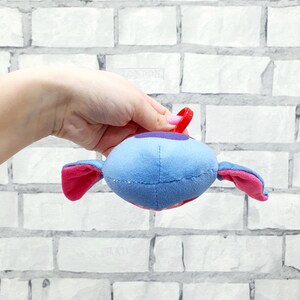 Może przedstawiać: Niebieska pluszowa zabawka w kształcie kosmity z r&oacute;żowymi uszami i czerwoną pętlą. Kosmita ma fioletowy pasek i czerwone usta. Zabawka jest trzymana na tle białej ceglanej ściany.