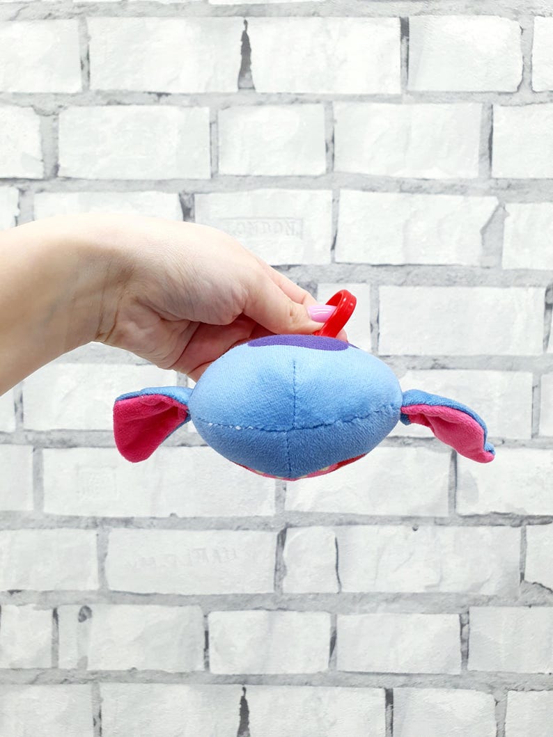 Może przedstawiać: Niebieska pluszowa zabawka w kształcie kosmity z r&oacute;żowymi uszami i czerwoną pętlą. Kosmita ma fioletowy pasek i czerwone usta. Zabawka jest trzymana na tle białej ceglanej ściany.