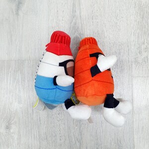 May include: Two plush toys shaped like cartoon characters. One is white, blue, and red, with black glasses. The other is orange. Both have white hands and feet, and black accents. They are on a light wood-grain surface.