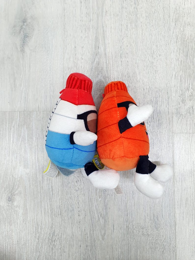 May include: Two plush toys shaped like cartoon characters. One is white, blue, and red, with black glasses. The other is orange. Both have white hands and feet, and black accents. They are on a light wood-grain surface.