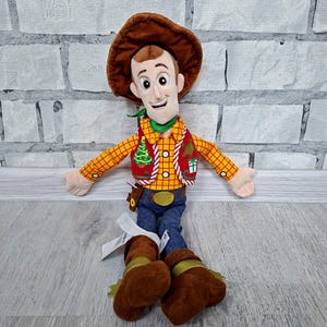 May include: A soft toy of Woody from Toy Story. The doll has a brown cowboy hat, a yellow plaid shirt, a red waistcoat, blue jeans, and brown boots. It has a green neckerchief and a smiling face. The toy is designed to resemble the character from the animated film.