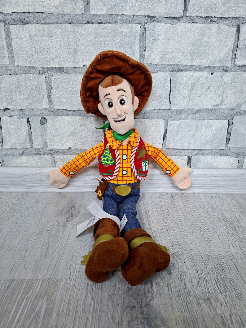 May include: A soft toy of Woody from Toy Story. The doll has a brown cowboy hat, a yellow plaid shirt, a red waistcoat, blue jeans, and brown boots. It has a green neckerchief and a smiling face. The toy is designed to resemble the character from the animated film.