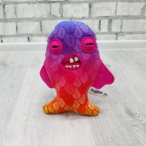May include: A plush toy in the shape of a colorful fish-like creature. The toy features a gradient of colors, including purple, pink, red, and orange. It has large, round eyes, a wide mouth with teeth, and small fins. The toy is soft and cuddly.