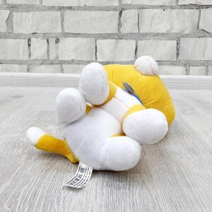 May include: A plush toy cat in a playful pose, lying on its back with its paws in the air. The cat features a white body with yellow accents on its head, tail, and paws. The toy has a tag attached.