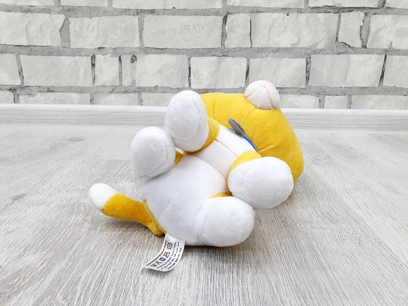May include: A plush toy cat in a playful pose, lying on its back with its paws in the air. The cat features a white body with yellow accents on its head, tail, and paws. The toy has a tag attached.
