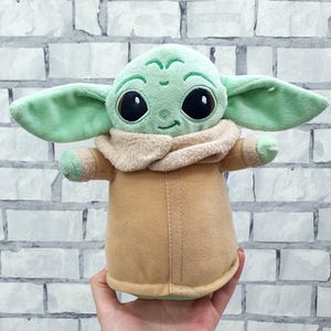May include: A plush doll of a character with large black eyes, green skin, and large ears. The doll is wearing a tan coat with a beige collar. The doll is being held in front of a white brick wall.