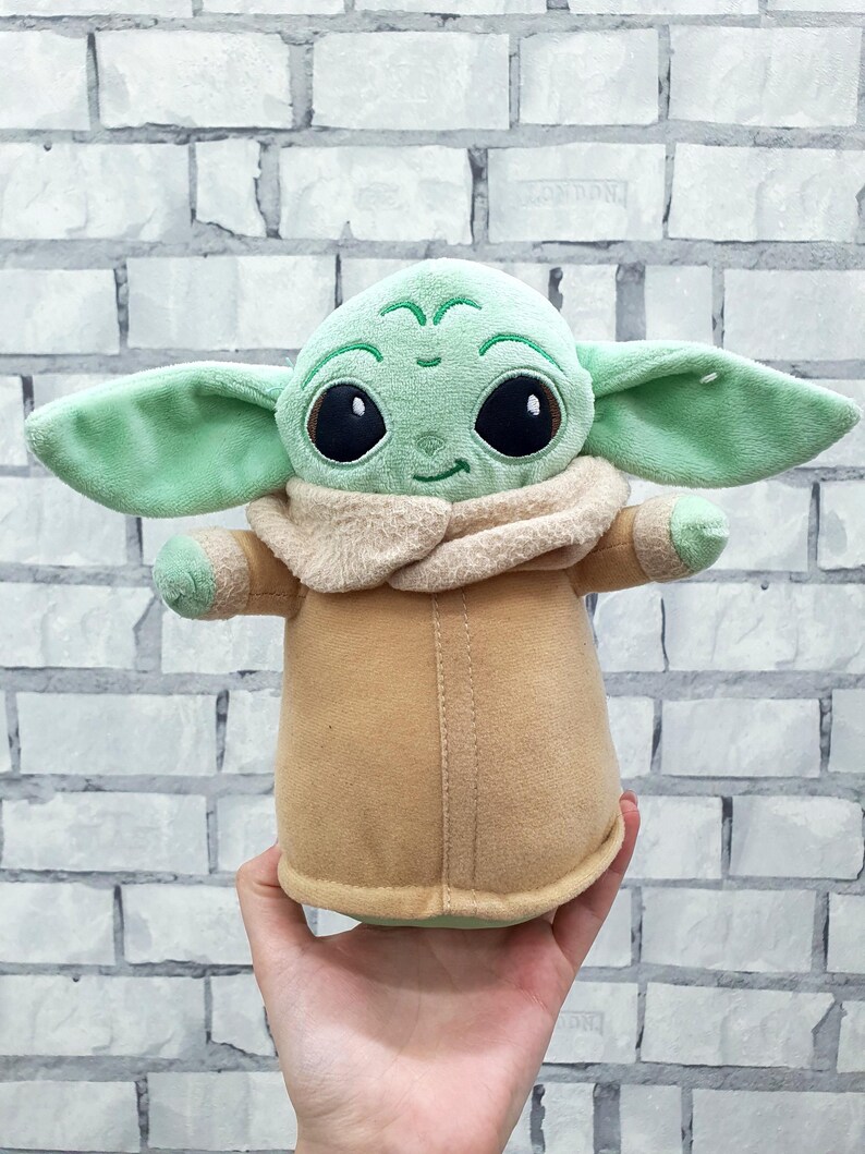 May include: A plush doll of a character with large black eyes, green skin, and large ears. The doll is wearing a tan coat with a beige collar. The doll is being held in front of a white brick wall.