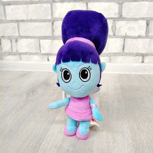 May include: A plush doll with a blue face, arms, and legs, and a purple bun. The doll is wearing a pink dress and shoes. The doll has large black eyes and a smiling expression. The doll is standing in front of a white brick wall.